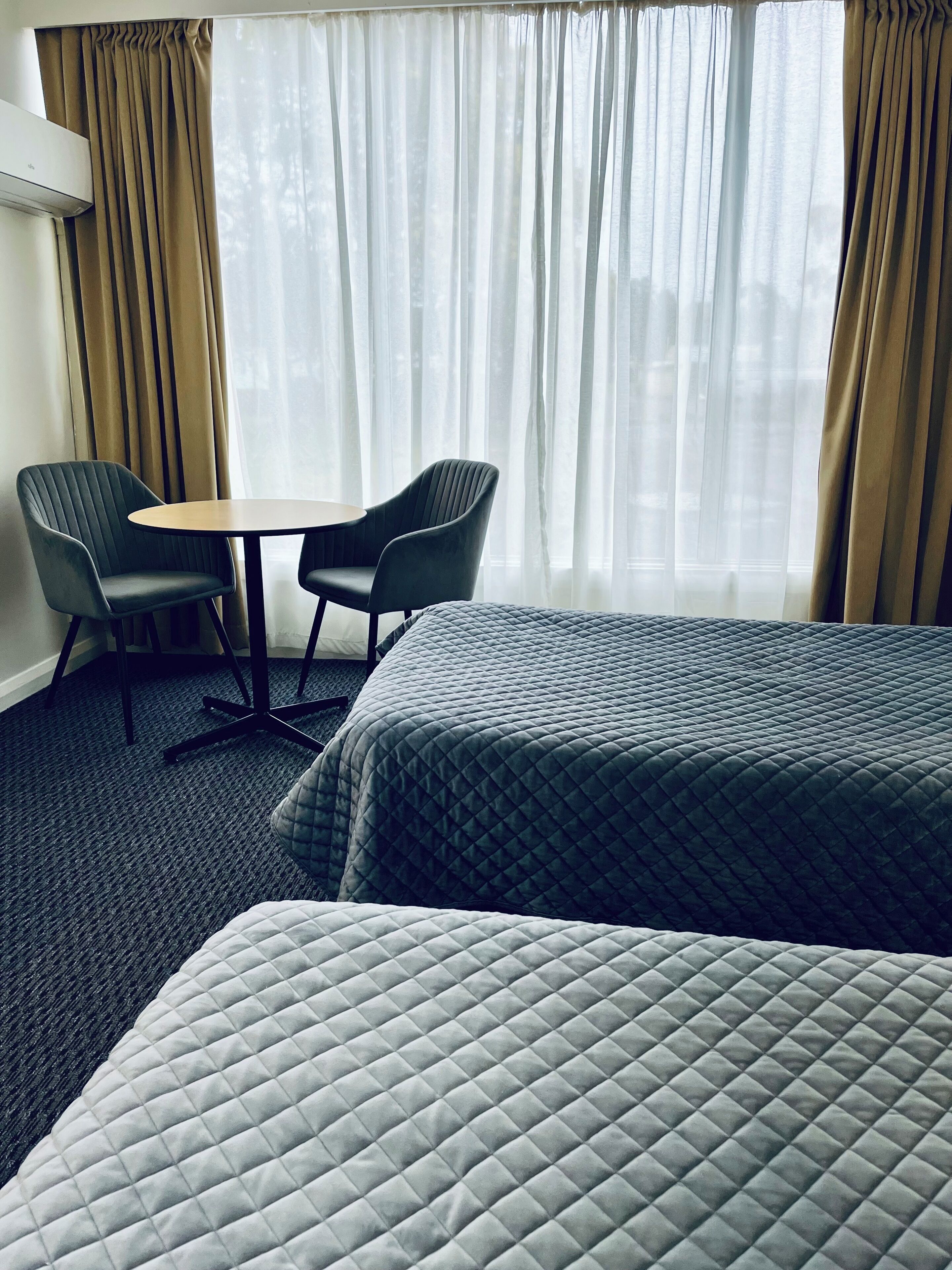 Una habitación de hotel con dos camas, una mesita pequeña, dos sillas y un ventanal con cortinas.