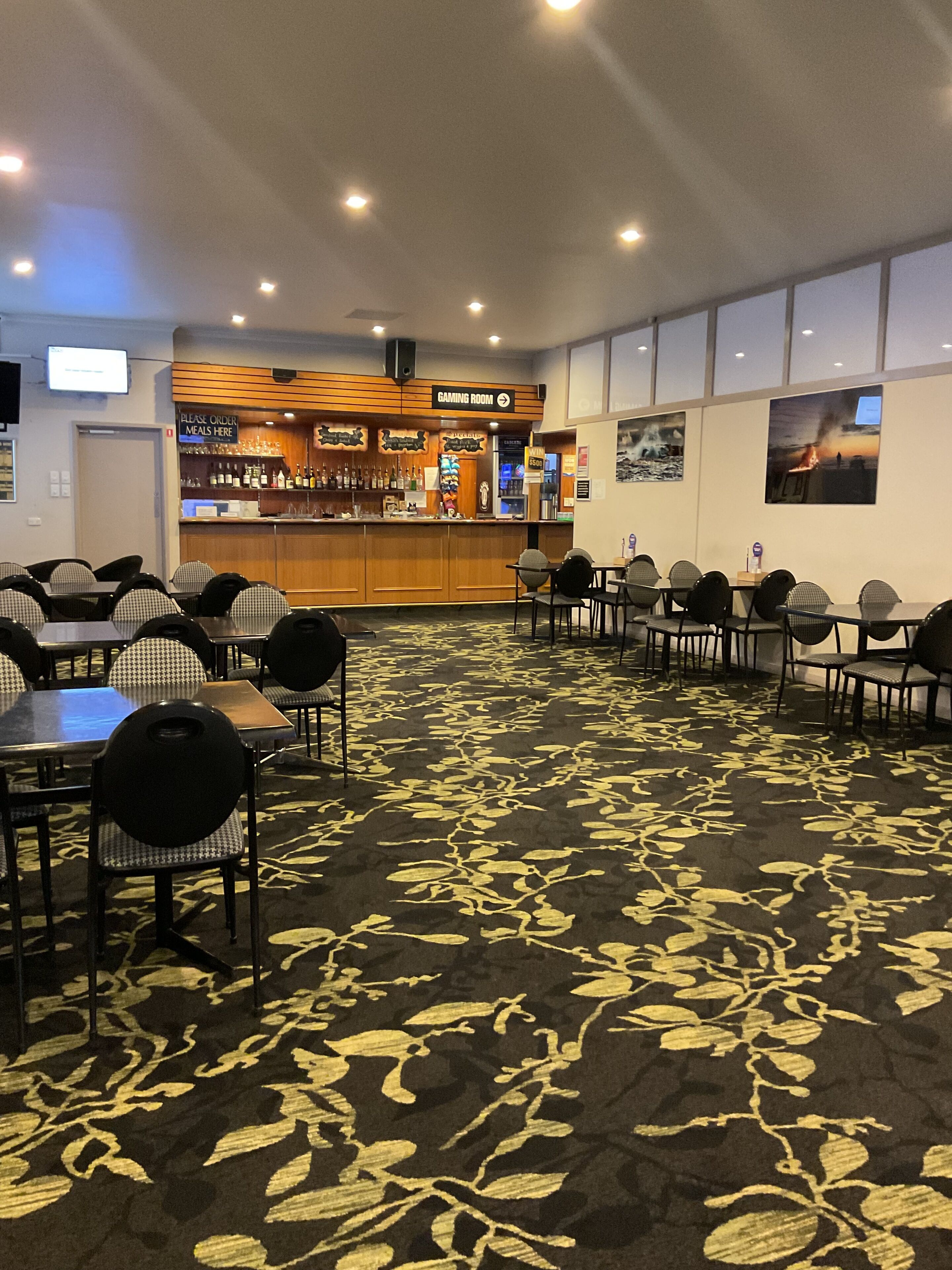 Un hall de hotel con zona de bar, varias mesas y sillas, y suelo alfombrado.