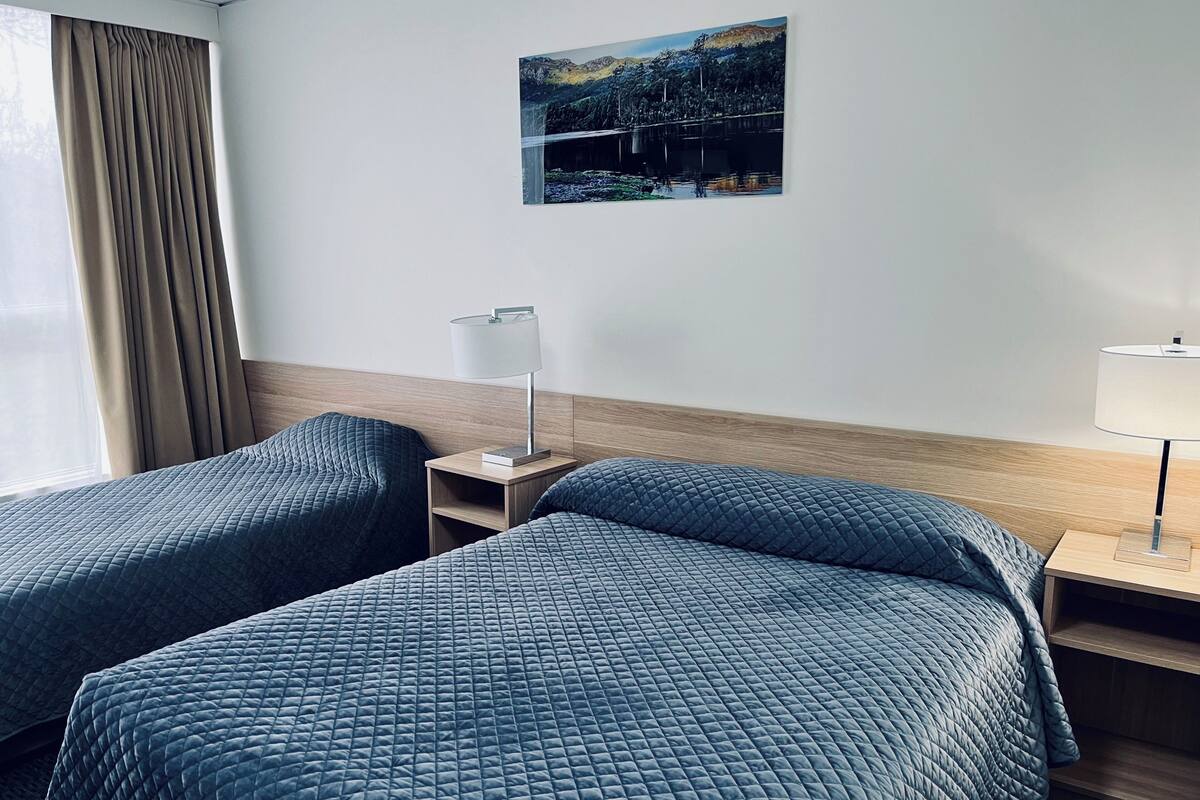 Une chambre d’hôtel avec deux lits, une table de chevet avec une lampe, un tableau au mur et une fenêtre avec des rideaux.