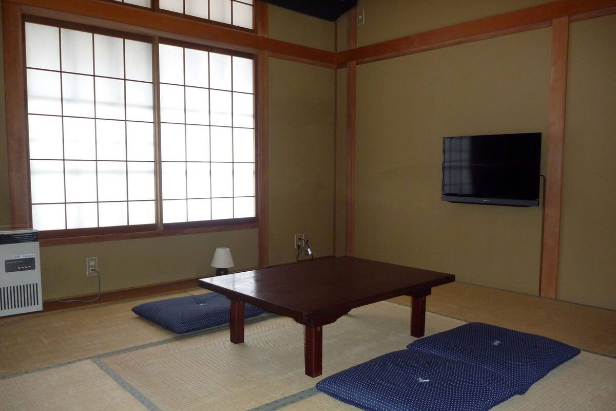 A traditional Japanese room with tatami mats, a low table, a heater, and a TV mounted on the wall.