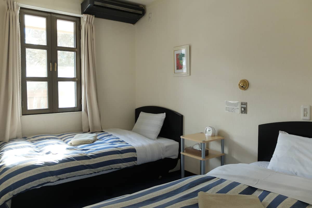 A hotel room with two beds, a window with curtains, a bedside table with a lamp, and a framed picture on the wall.
