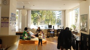 A spacious living room with a large window, a sofa, a child playing, and a dining area. - K's House Hakuba Alps - Hostel (Hakuba)