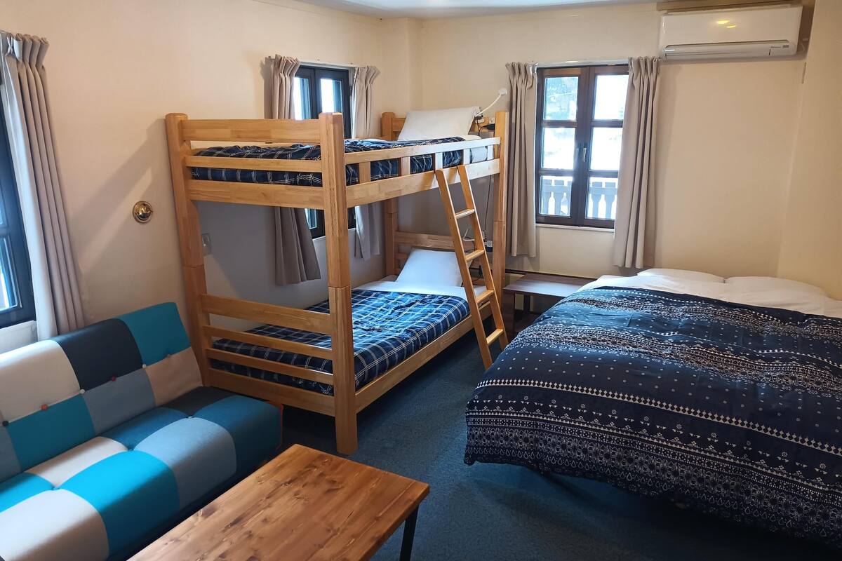 A bunk bed room with a wooden ladder, a sofa, a table, and a bed with patterned bedding.