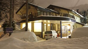 A two-story building with a balcony, a sign indicating it is a backpackers hostel, and a car partially buried in snow. - K's House Hakuba Alps - Hostel (Hakuba)