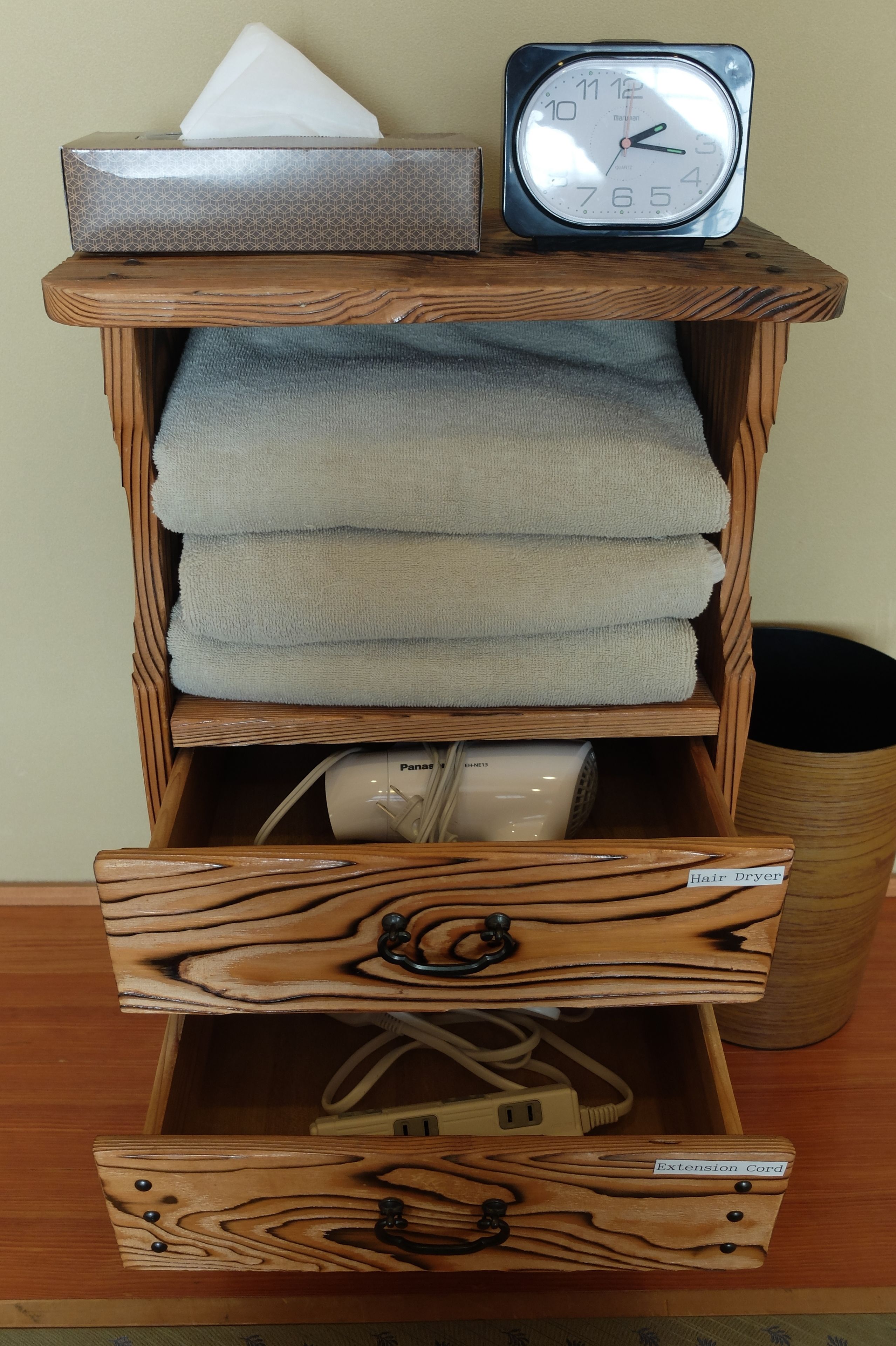 A wooden shelf with a tissue box, a clock, folded towels, and a hair dryer.