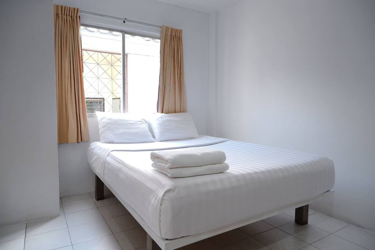 A neatly made bed with white linens and a folded blanket, a window with beige curtains, and a tiled floor.