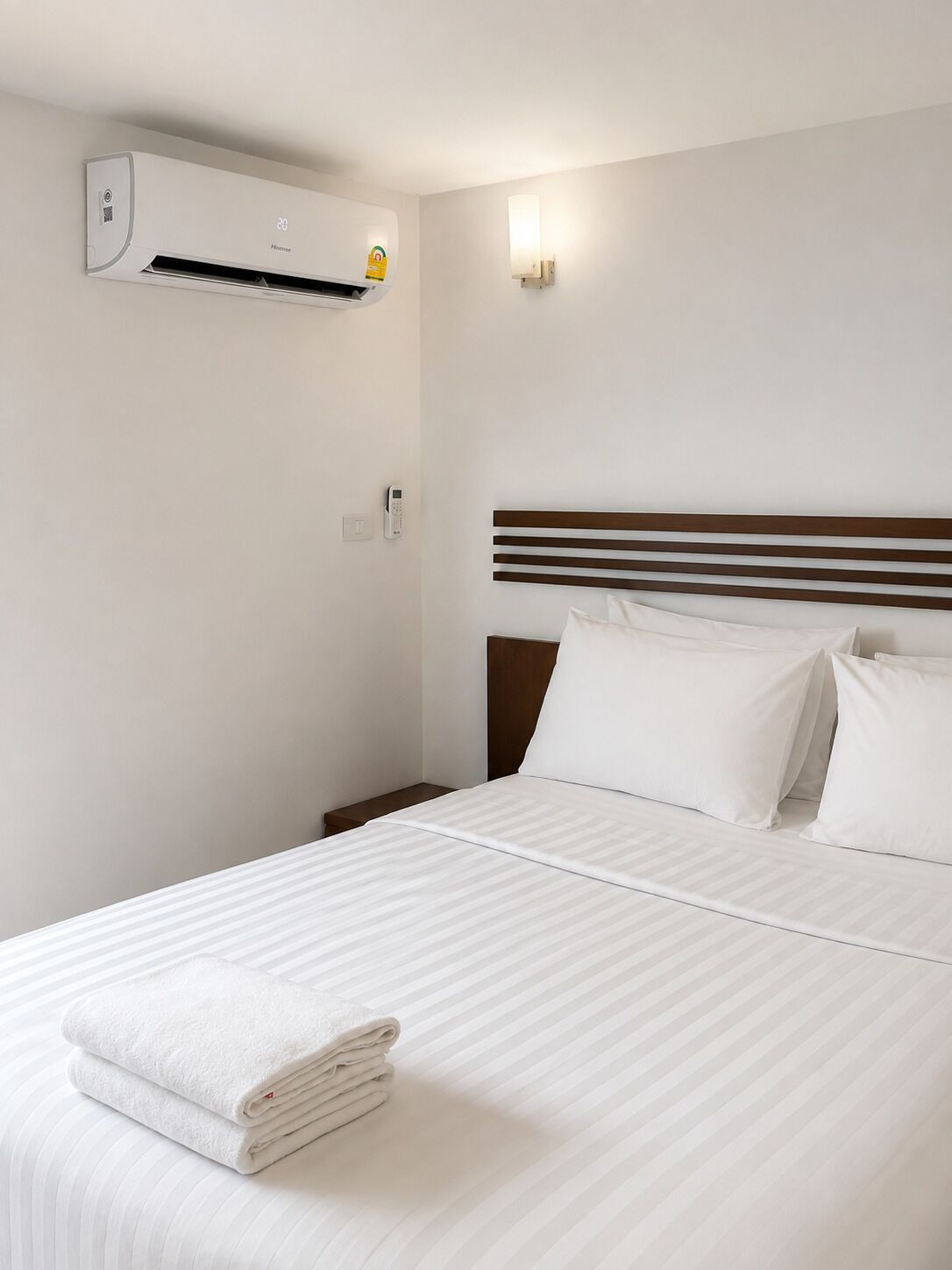 A hotel room with a bed, a wall-mounted air conditioner, a bedside table, and a wall-mounted light fixture.