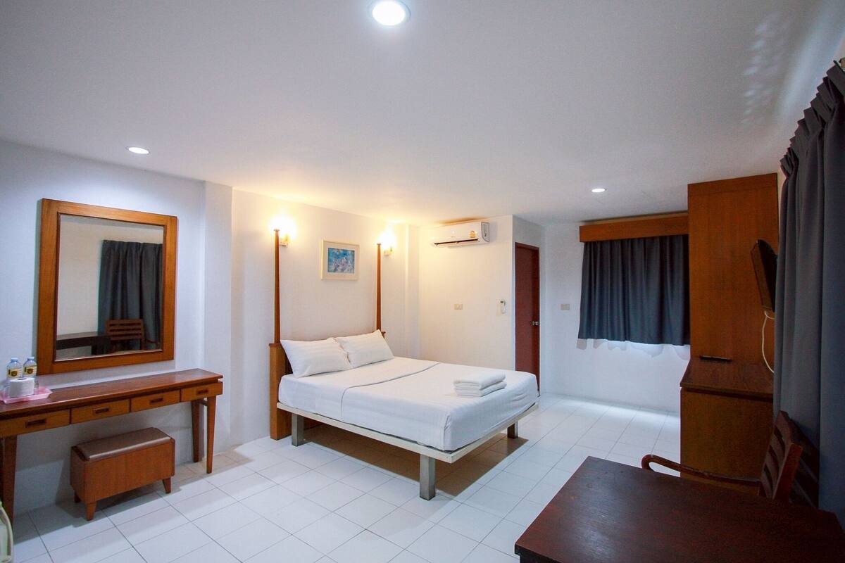 A hotel room with a bed, a desk, a chair, a small table, a mirror, and a window with curtains.