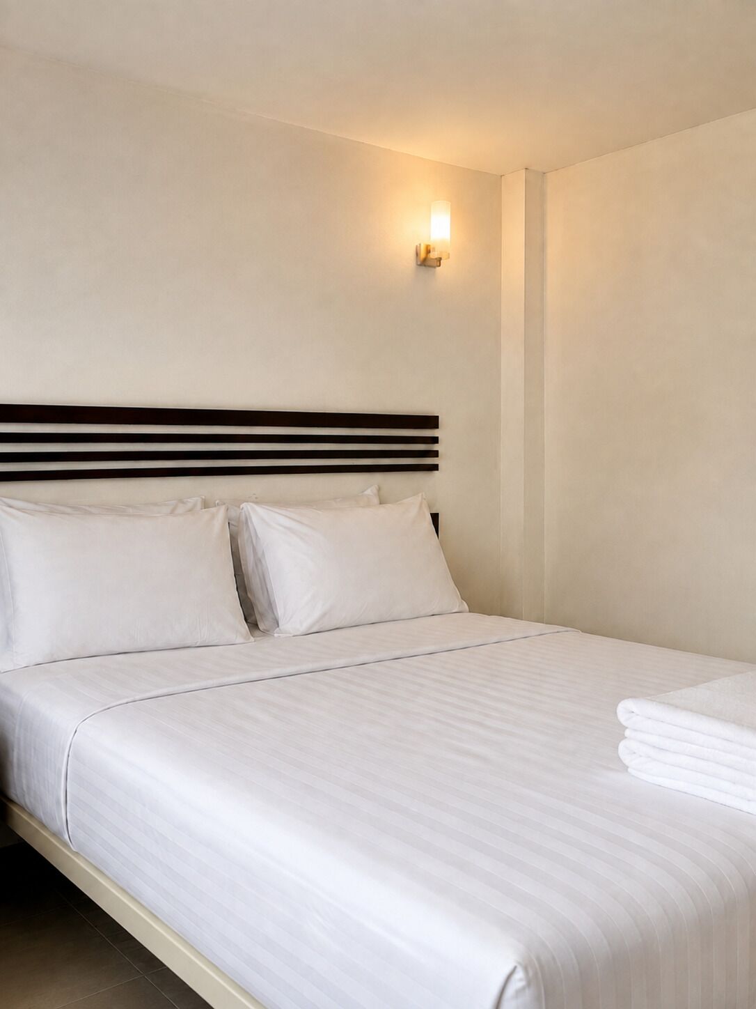 A neatly made bed with white linens and pillows, a dark headboard, and a wall-mounted light fixture.