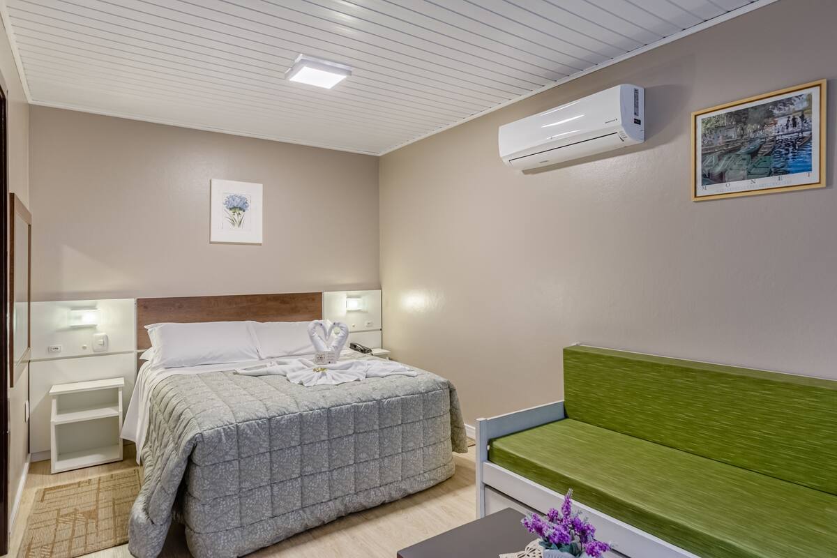 Quarto de hotel com cama, mesas de cabeceira, sofá verde, ar condicionado e um quadro na parede.