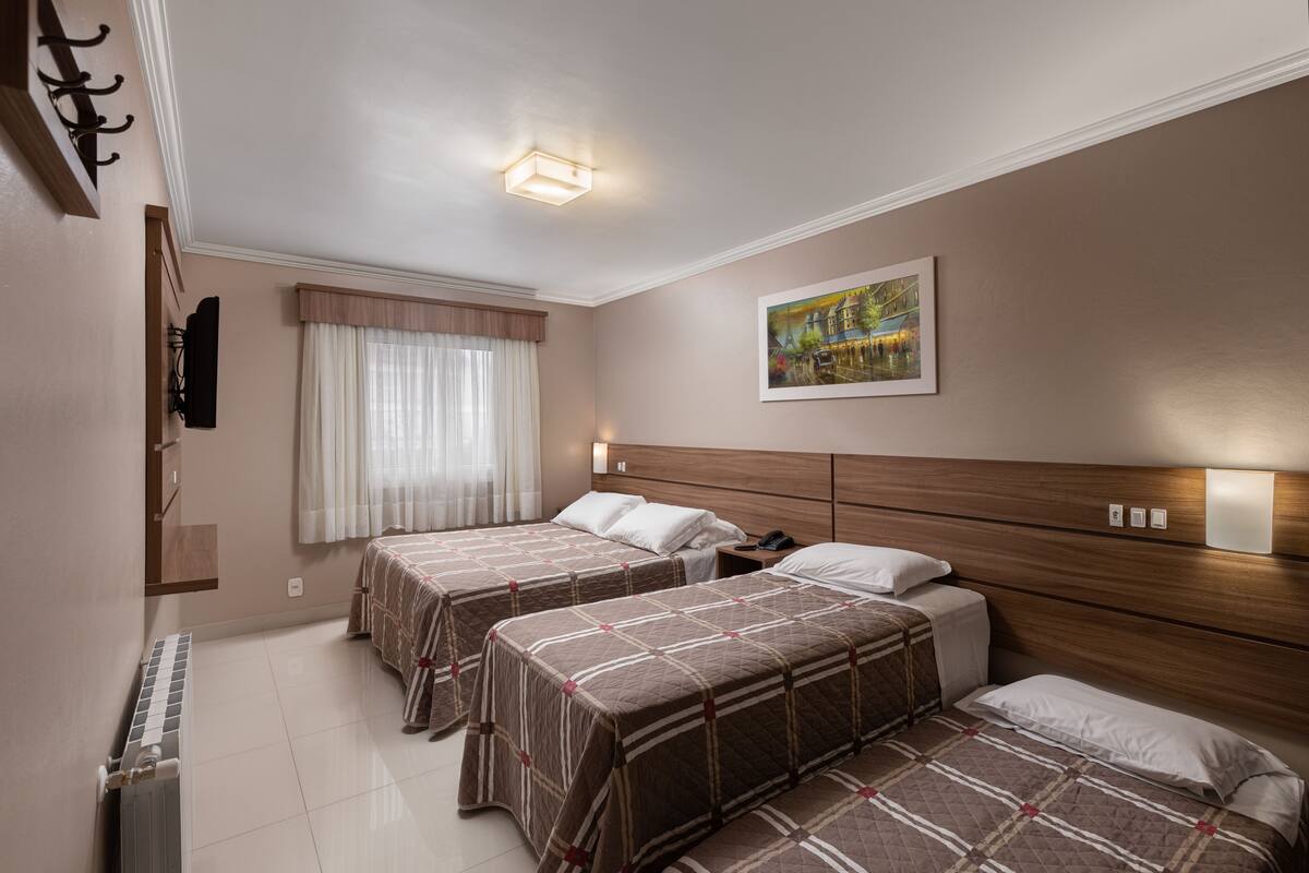 Quarto de hotel com duas camas, uma janela grande com cortinas leves, um quadro na parede e um radiador no chão.