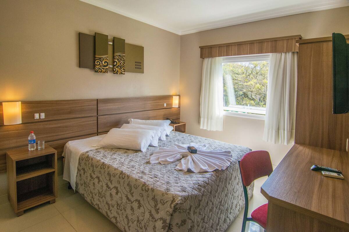 Quarto de hotel com cama, mesa de cabeceira, escrivaninha e cadeira, janela com cortinas e televisão na parede.
