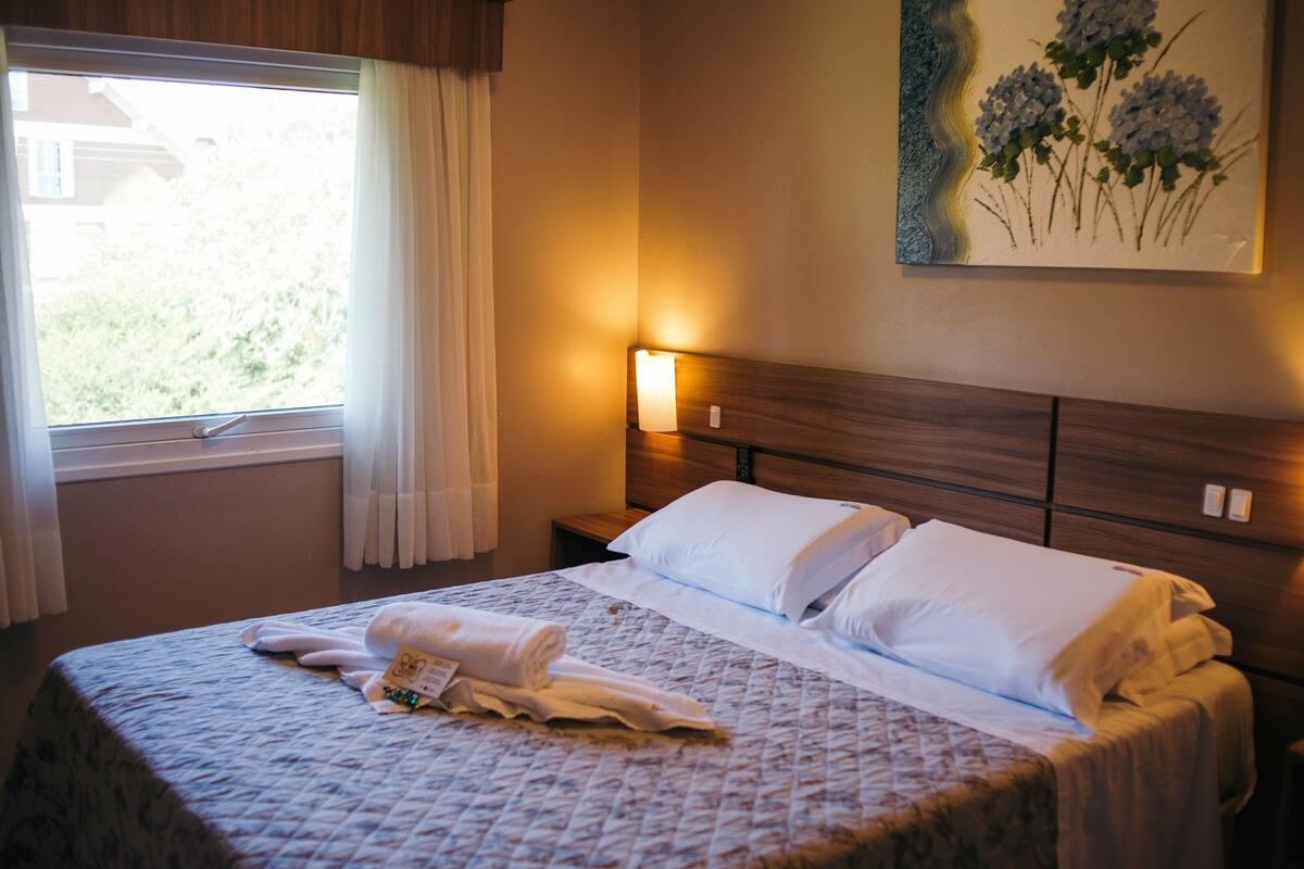 Uma cama bem arrumada com lençóis brancos e uma toalha enrolada, um abajur e um quadro de flores na parede.