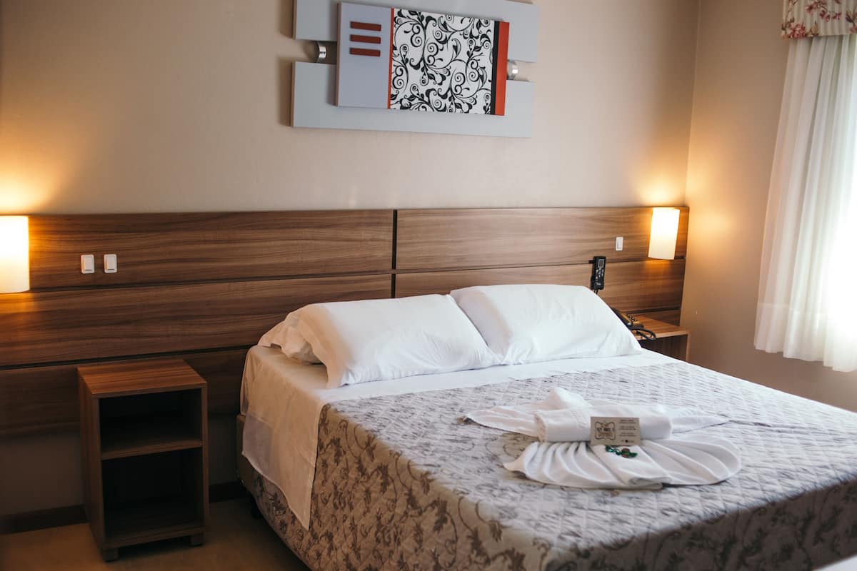 Quarto de hotel com cabeceira de madeira, cama com roupa de cama branca, mesa de cabeceira e uma obra de arte pendurada na parede.