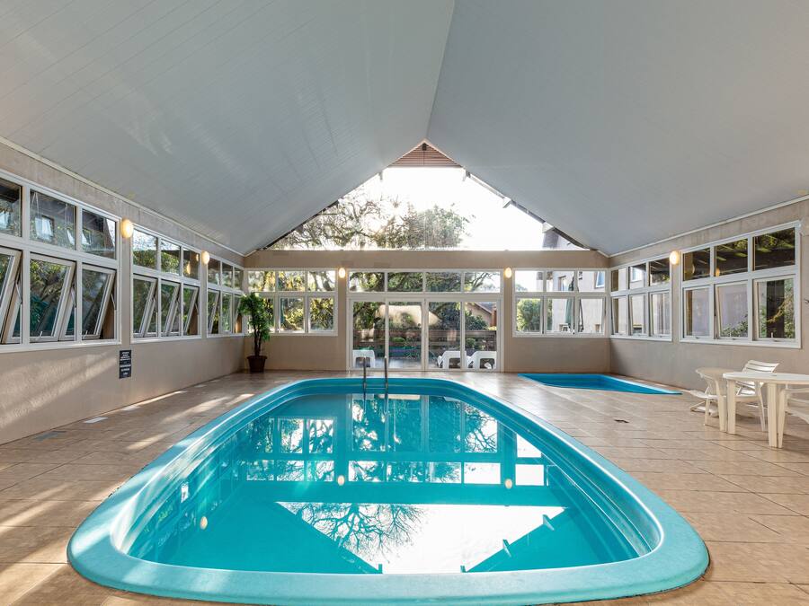 An indoor swimming pool with a clear view of the outside through large windows, a tiled floor, and a few white plastic chairs.