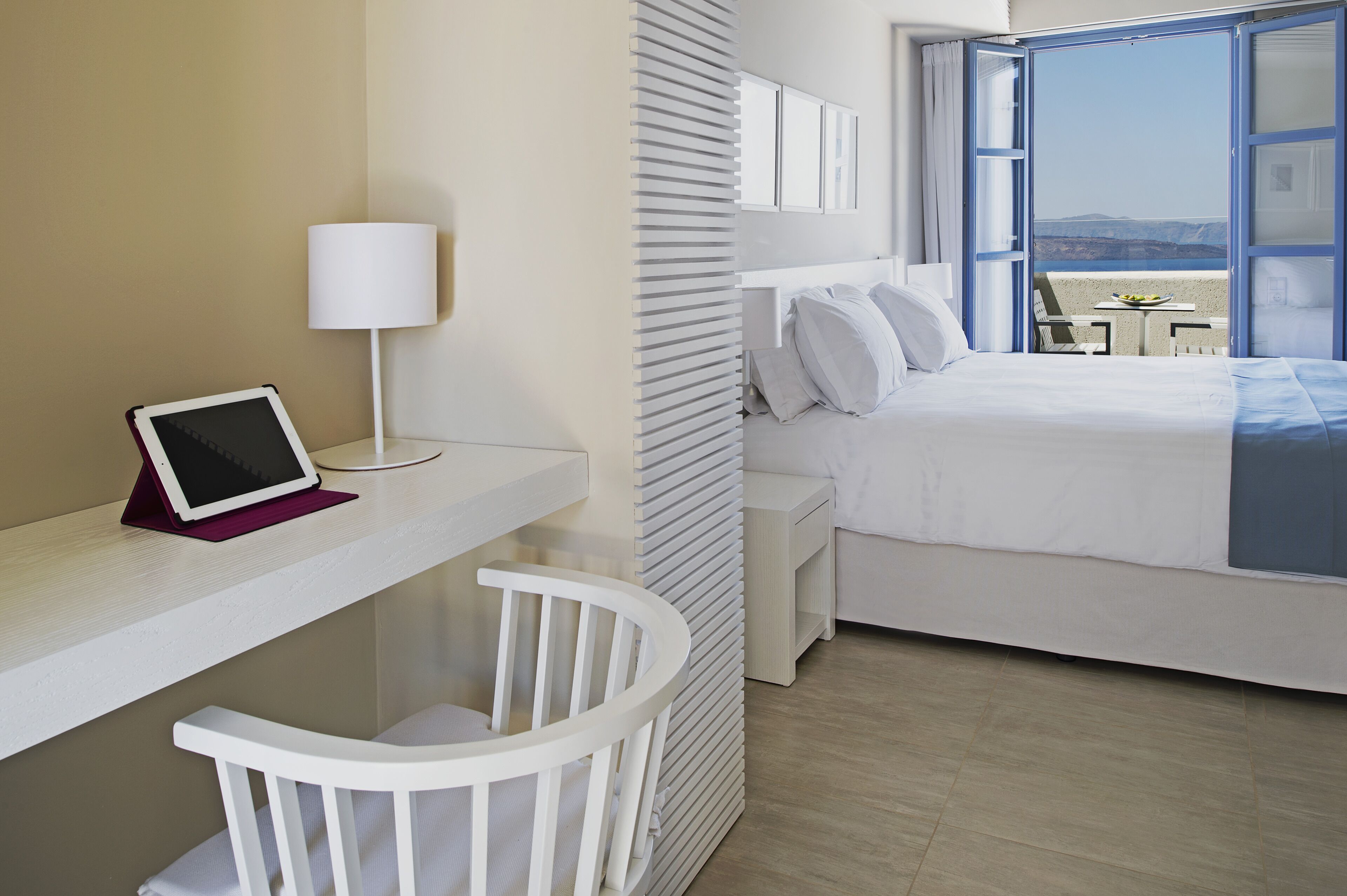 A hotel room with a bed, a desk with a tablet, a chair, a lamp, and a view of the outdoors.