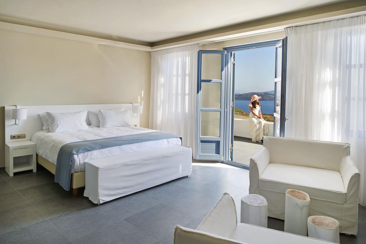 A hotel room with a bed, a sofa, a small table, and a view of the sea through a glass door.