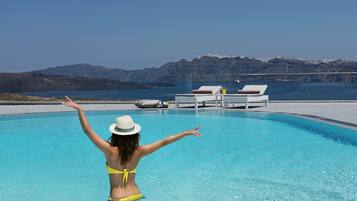 Une personne vêtue d’un bikini jaune et d’un chapeau blanc se tient dans une piscine, les bras levés, et surplombe un paysage montagneux sous un ciel bleu clair.