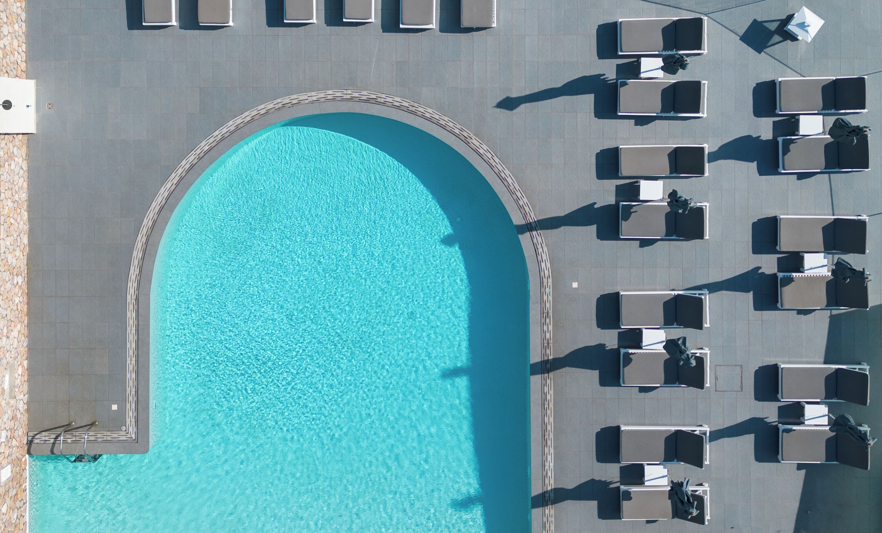 An aerial view of a swimming pool with a tiled deck, surrounded by lounge chairs and umbrellas.