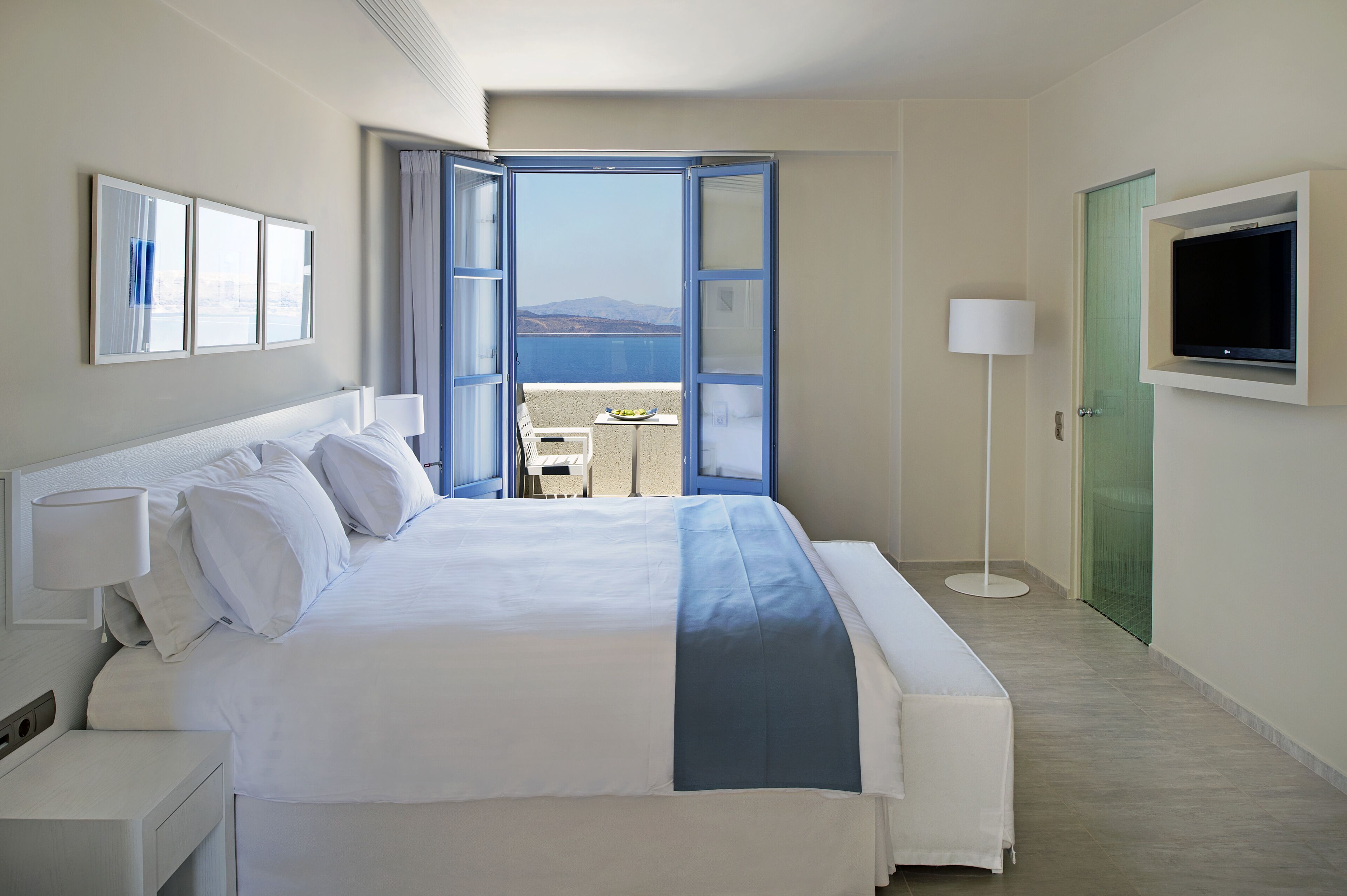 Een hotelkamer met een bed, een televisie, een lamp en uitzicht op zee door een geopende deur.