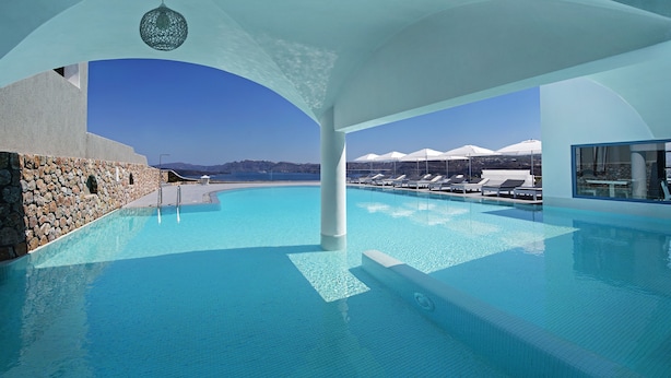 Una amplia piscina interior con agua cristalina, rodeada de paredes blancas y un muro de piedra en un lateral.