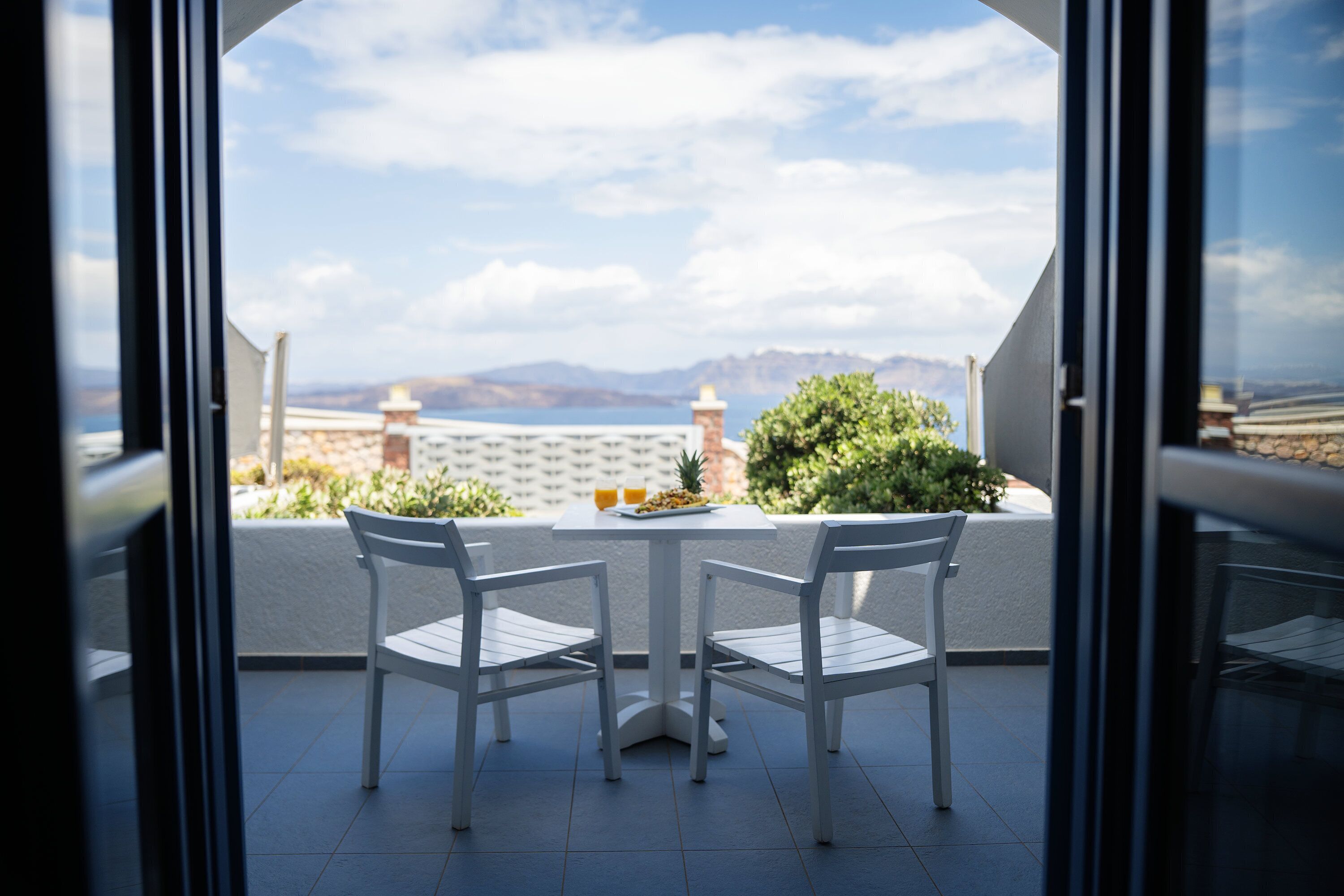 A balcony with two white chairs, a small table, and a view of the sea and mountains.
