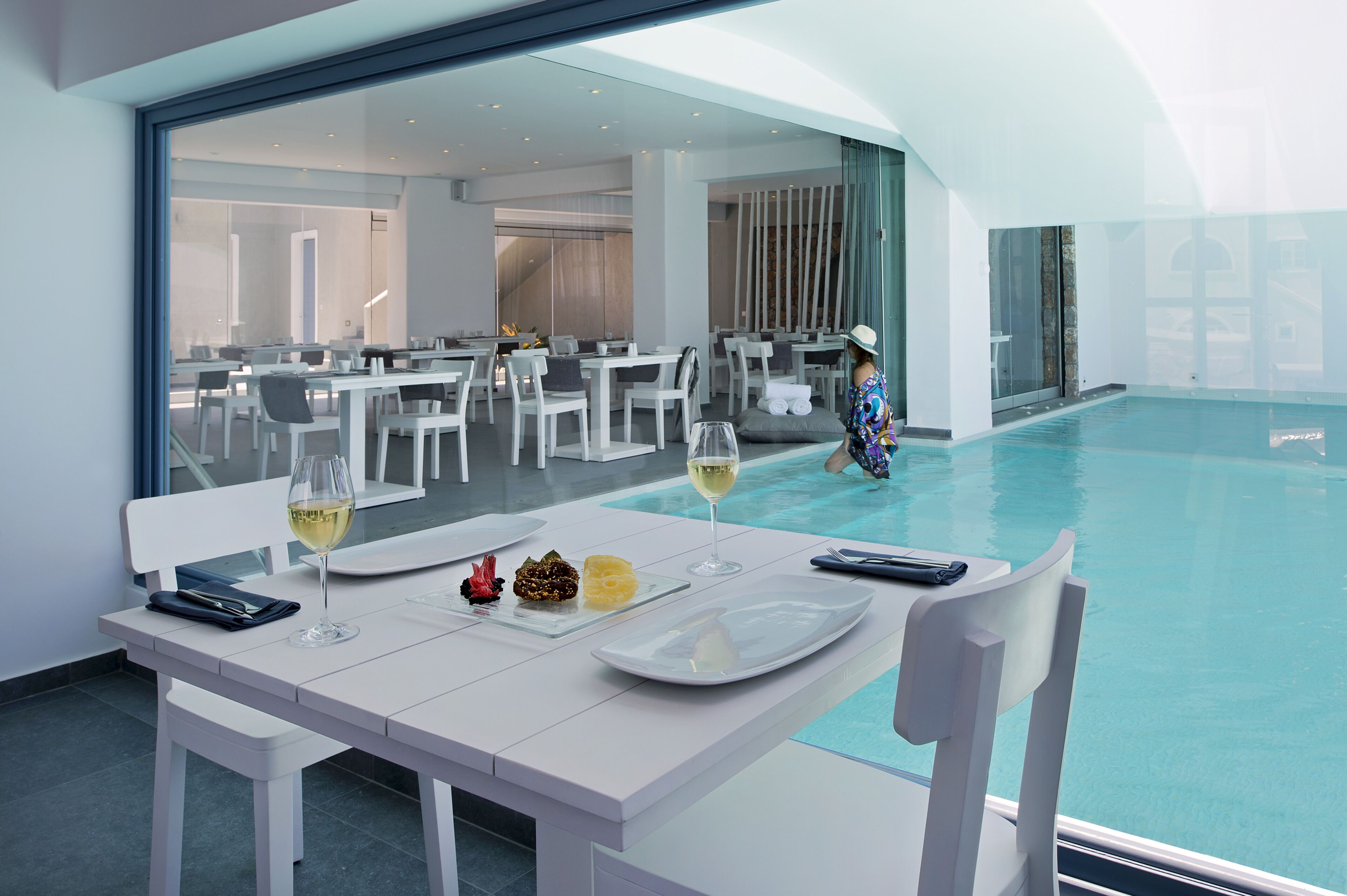 A dining area with white tables and chairs, a pool, and a person in a hat walking by.