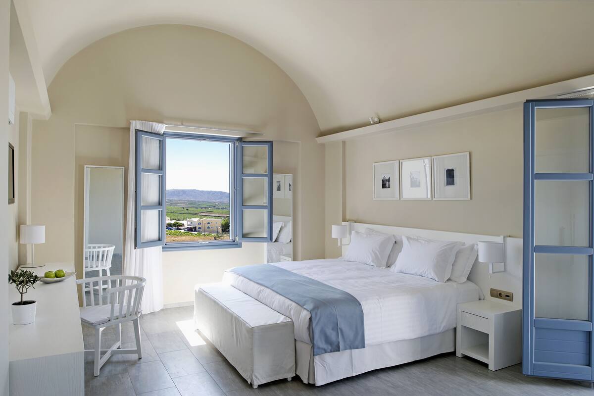 A bedroom with a large bed, a desk with a chair, a window with an open shutter, and a view of the countryside.