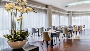 Un comedor espacioso con varias mesas y sillas, una orquídea en maceta grande y ventanas amplias con cortinas traslúcidas.