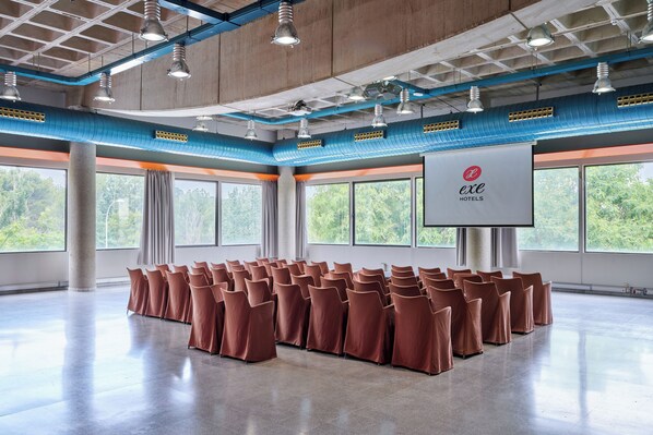 Un salón de conferencias con filas de sillas, una pantalla de proyector y grandes ventanales con vistas a árboles.