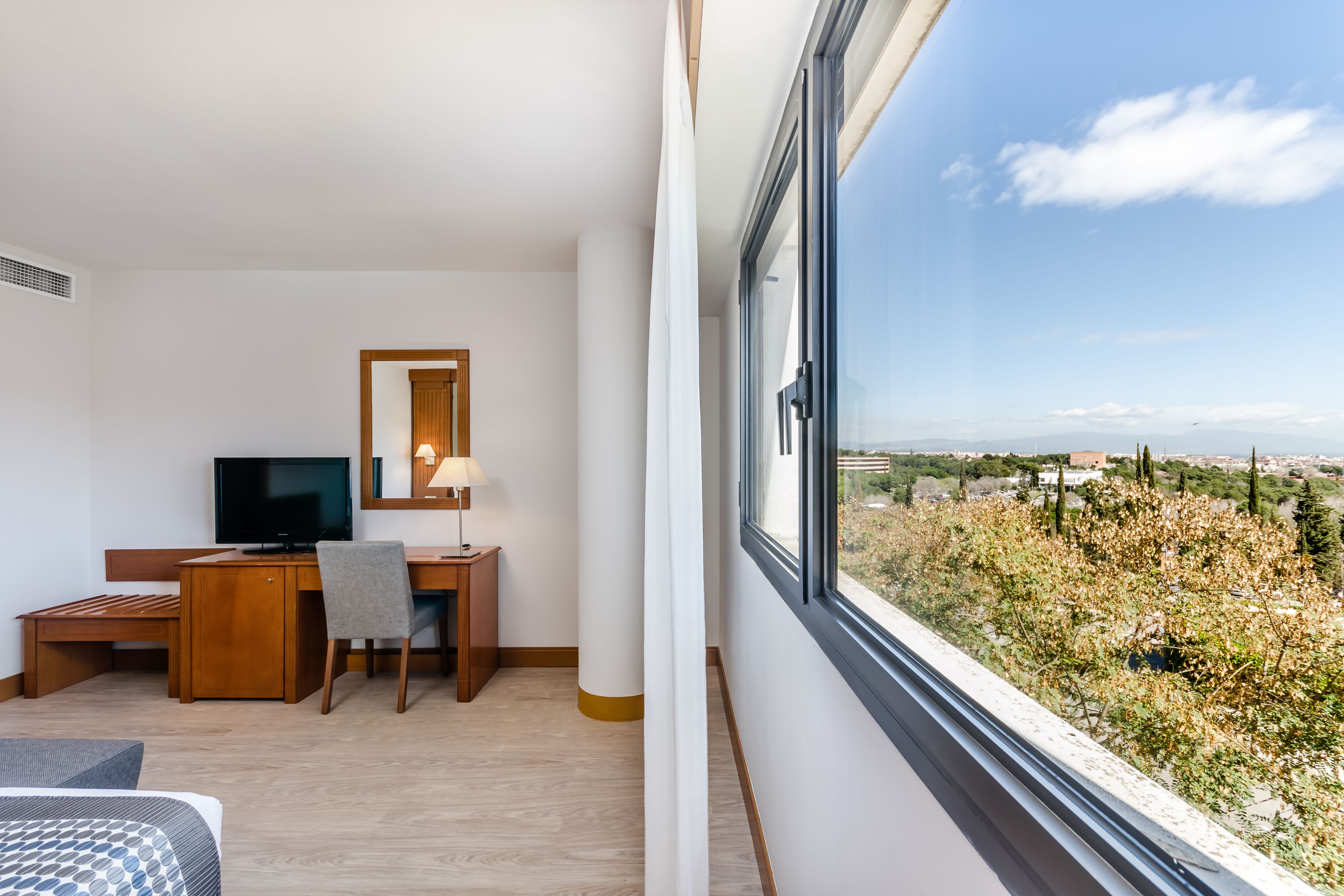 A hotel room with a desk, chair, television, and a large window offering a view of the countryside.