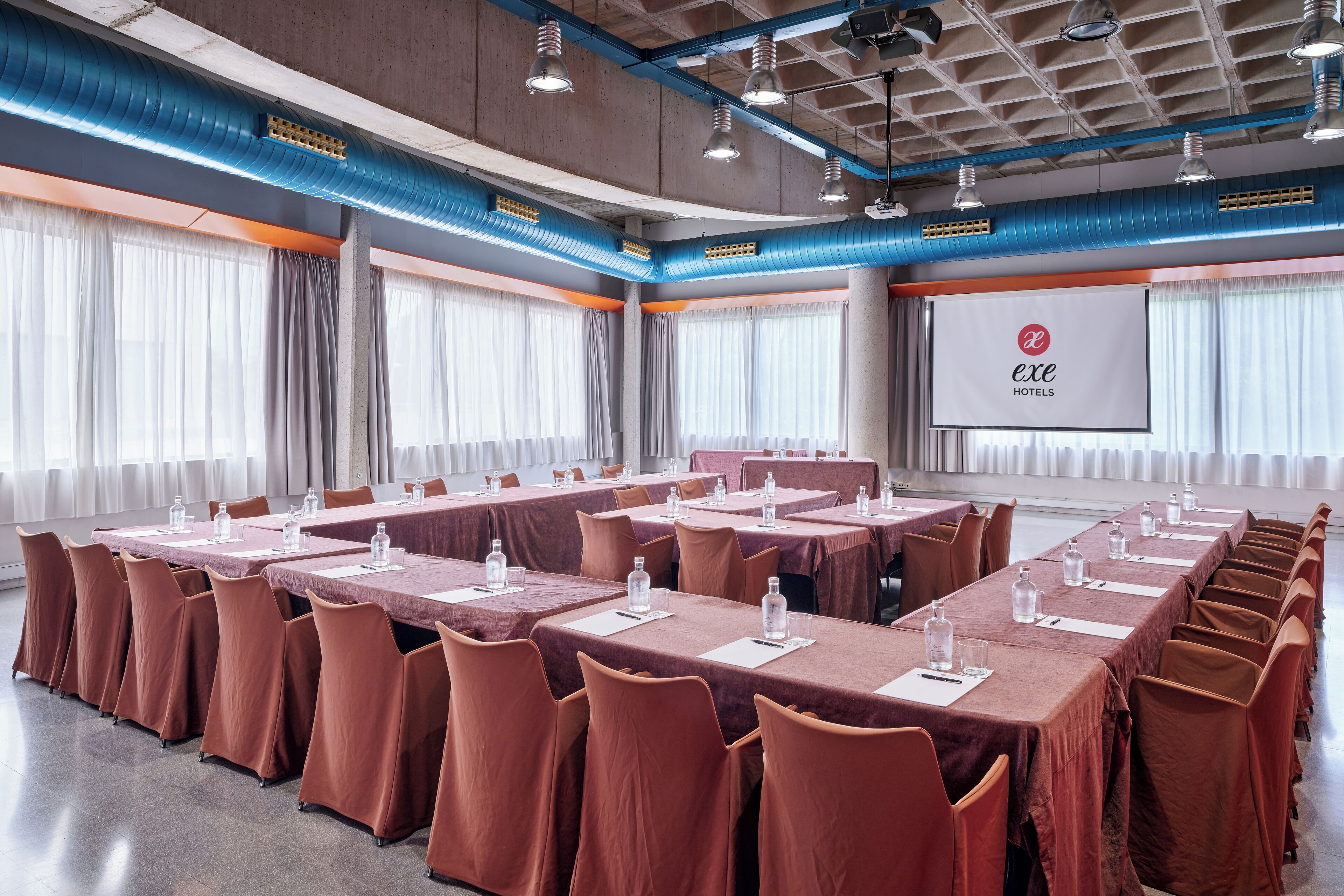 A conference room with long tables, chairs, and a projection screen displaying the logo of exe Hotels.