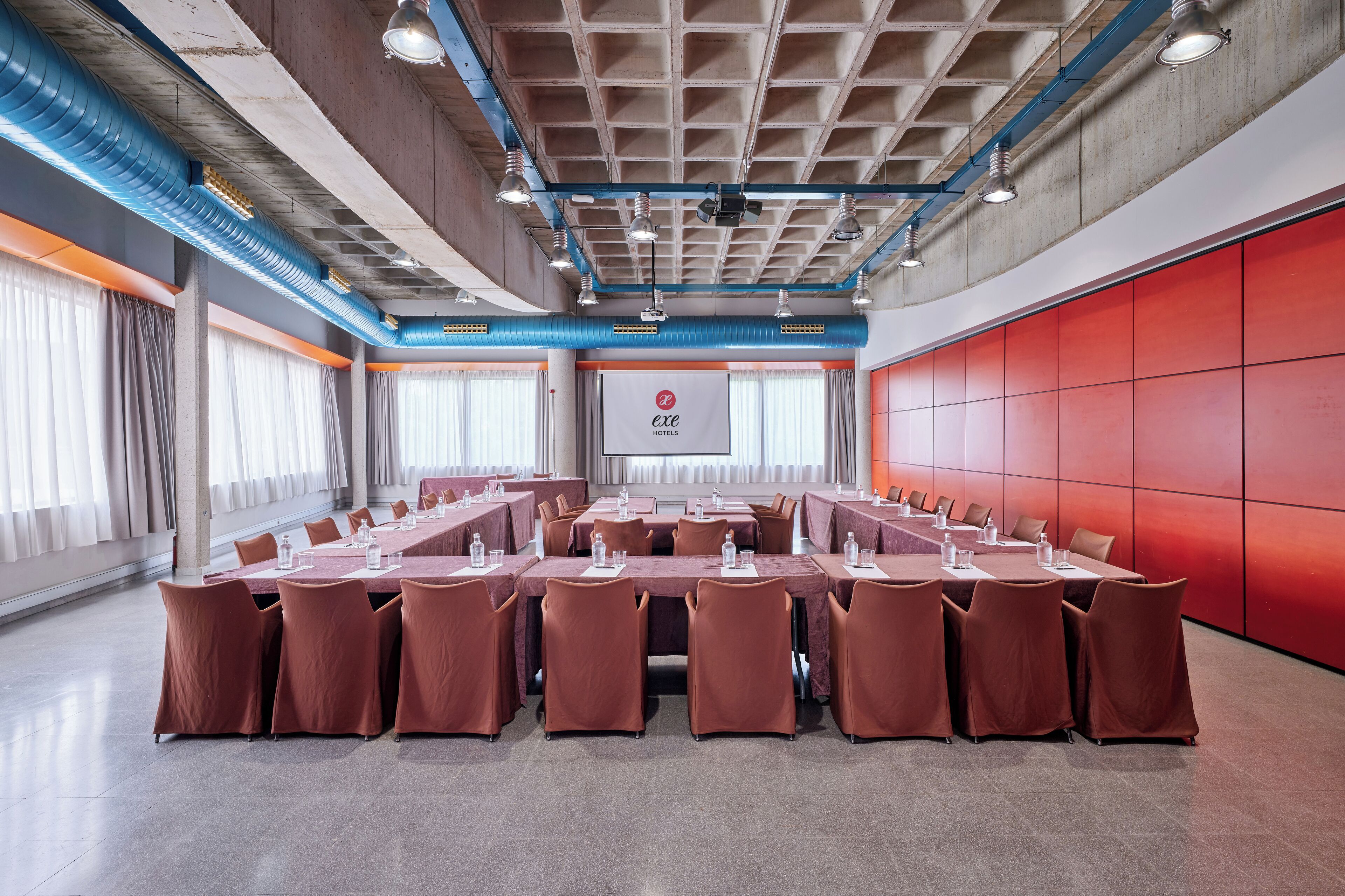 Un salón de conferencias con una larga mesa, múltiples sillas y una pared roja decorativa.
