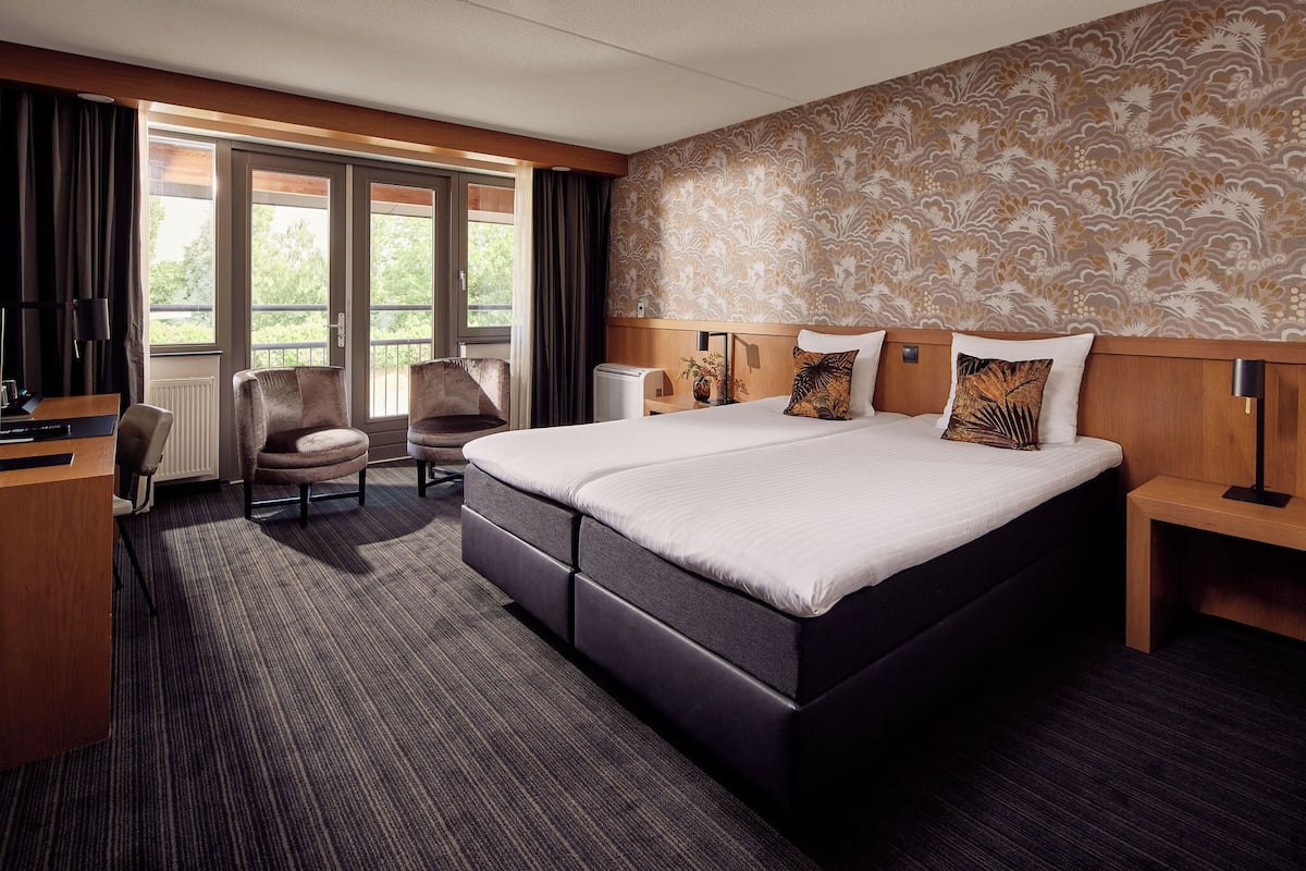 A hotel room with a large bed, a desk, a chair, and a view of greenery through the windows.