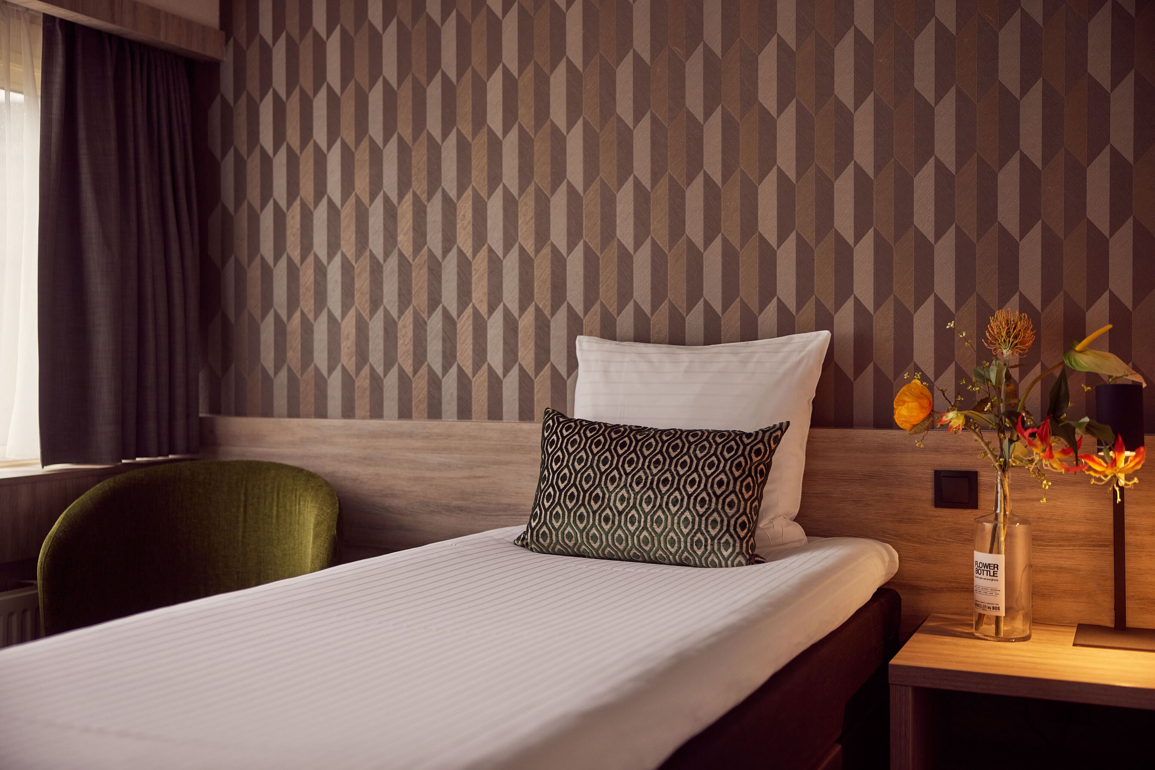 A hotel room with a neatly made bed, a green chair, a nightstand with a vase of flowers, and a patterned wall.