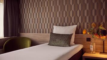 A hotel room with a neatly made bed, a green chair, a nightstand with a vase of flowers, and a patterned wall.