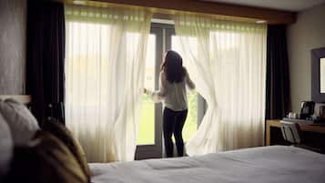 A hotel room with a bed, a desk with a chair, and a window with sheer curtains.
