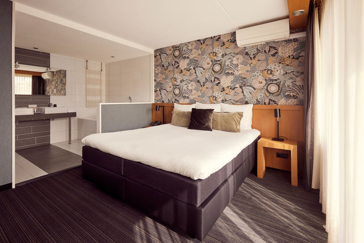 A modern hotel room with a large bed, a small bedside table, a nightstand, a built-in wardrobe, and a bathroom with a shower and sink.