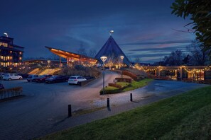 Un edificio moderno con un techo triangular distintivo, un estacionamiento con varios coches y una entrada bien iluminada.
