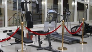 A gym with exercise equipment, including a treadmill and weight machines, enclosed by a glass wall with a red rope barrier. - Leonardo (Durres)
