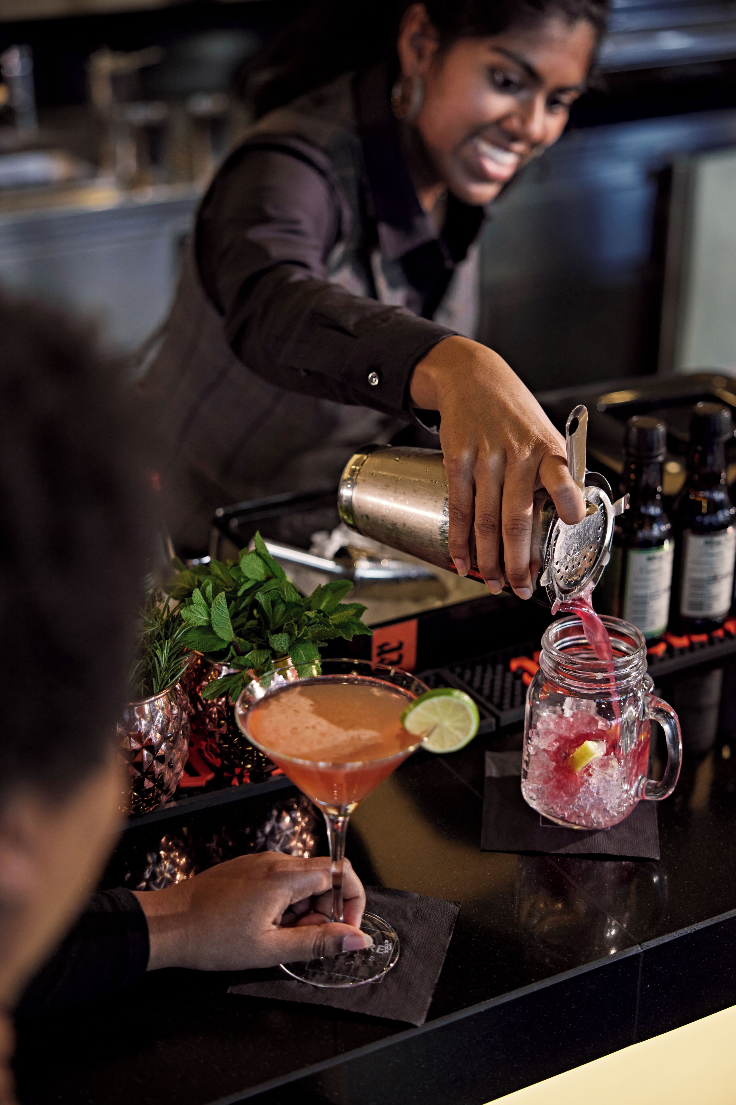 A bartender pouring a drink into a glass, with a lime wedge and mint leaves visible.