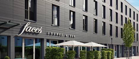 Un edificio moderno con un letrero que dice 'Lounge & Bar', asientos exteriores con sombrillas y macetas ordenadas.