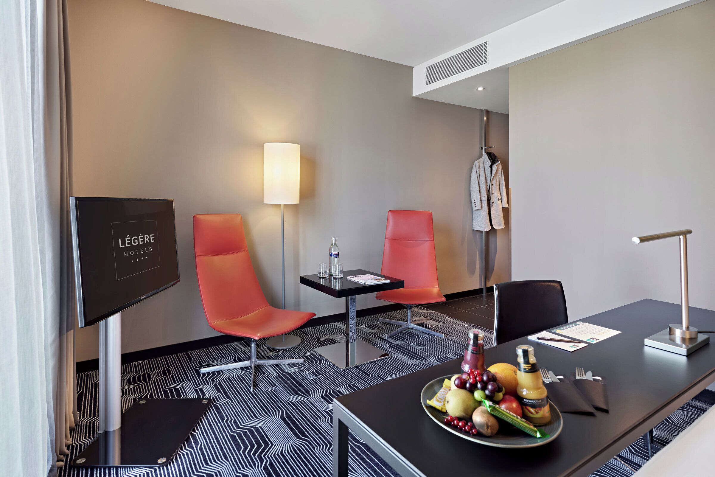 Quarto de hotel com um piso estampado em preto e branco, duas cadeiras vermelhas, uma mesa preta com um prato de frutas, uma televisão e um cabideiro.