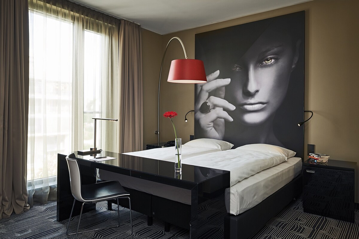 A hotel room with a bed, desk, chair, and a large wall art piece.