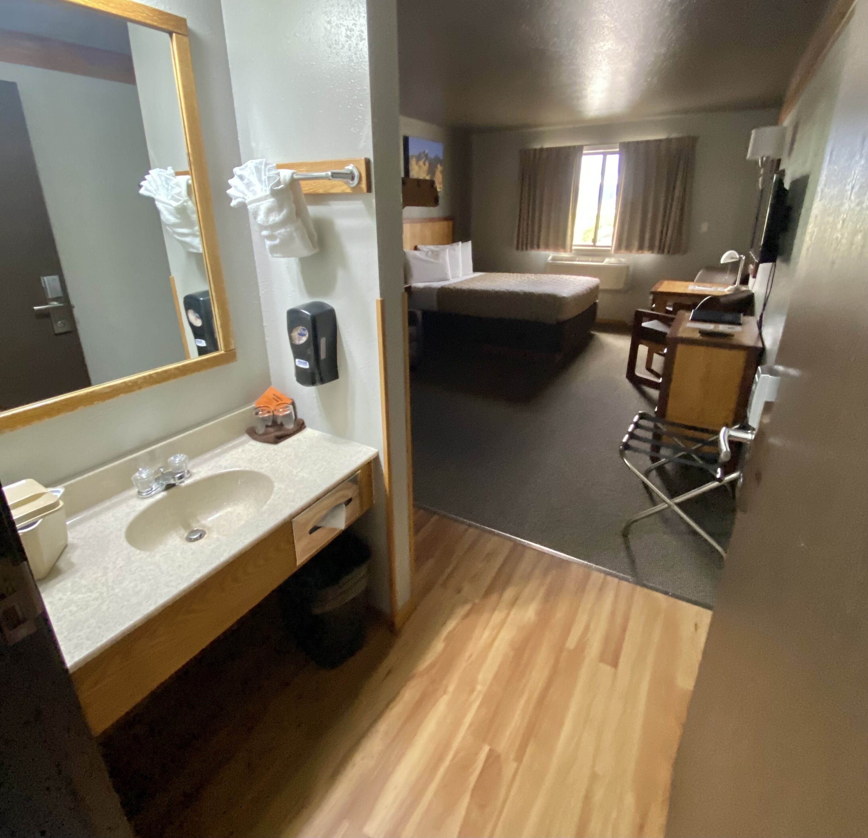 A hotel room with a wooden floor, a bed, a desk, a chair, a mirror, a sink, and a window.