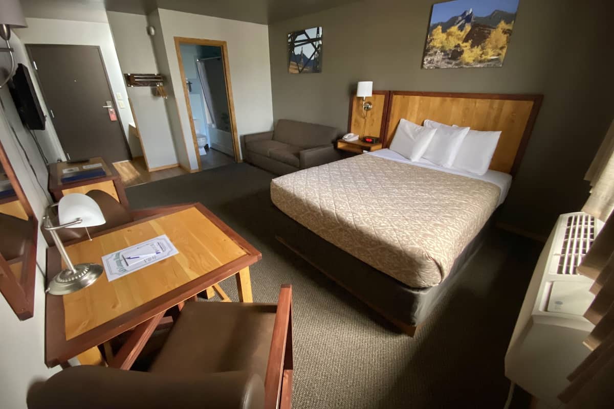 A hotel room with a bed, a desk with a chair, a sofa, a bedside table with a lamp, and a painting on the wall.