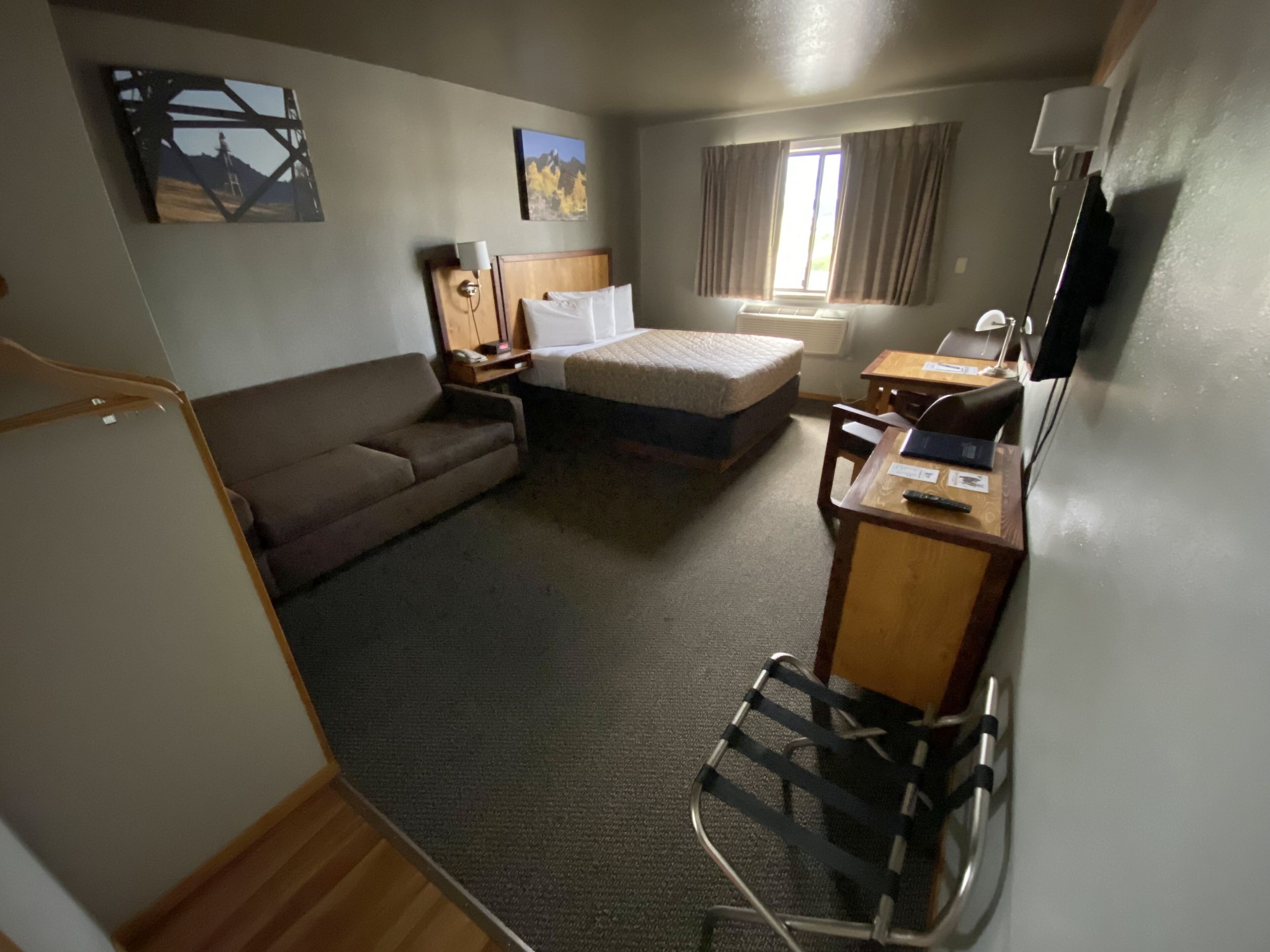 A compact hotel room with a bed, a desk, a chair, a sofa, a TV, and artwork on the walls.