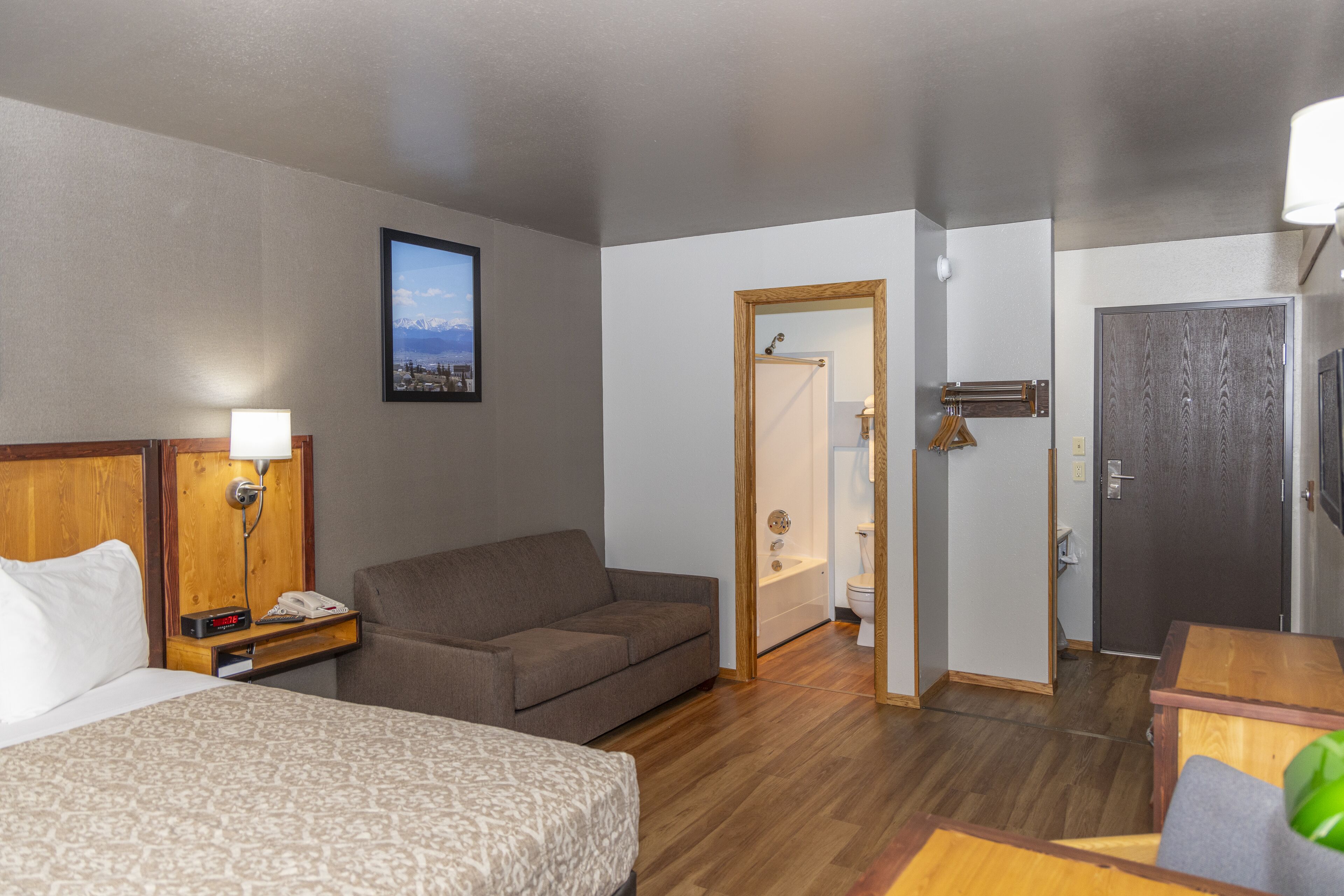 A hotel room with a bed, a sofa, a desk, a mirror, a picture on the wall, and a door leading to a bathroom.