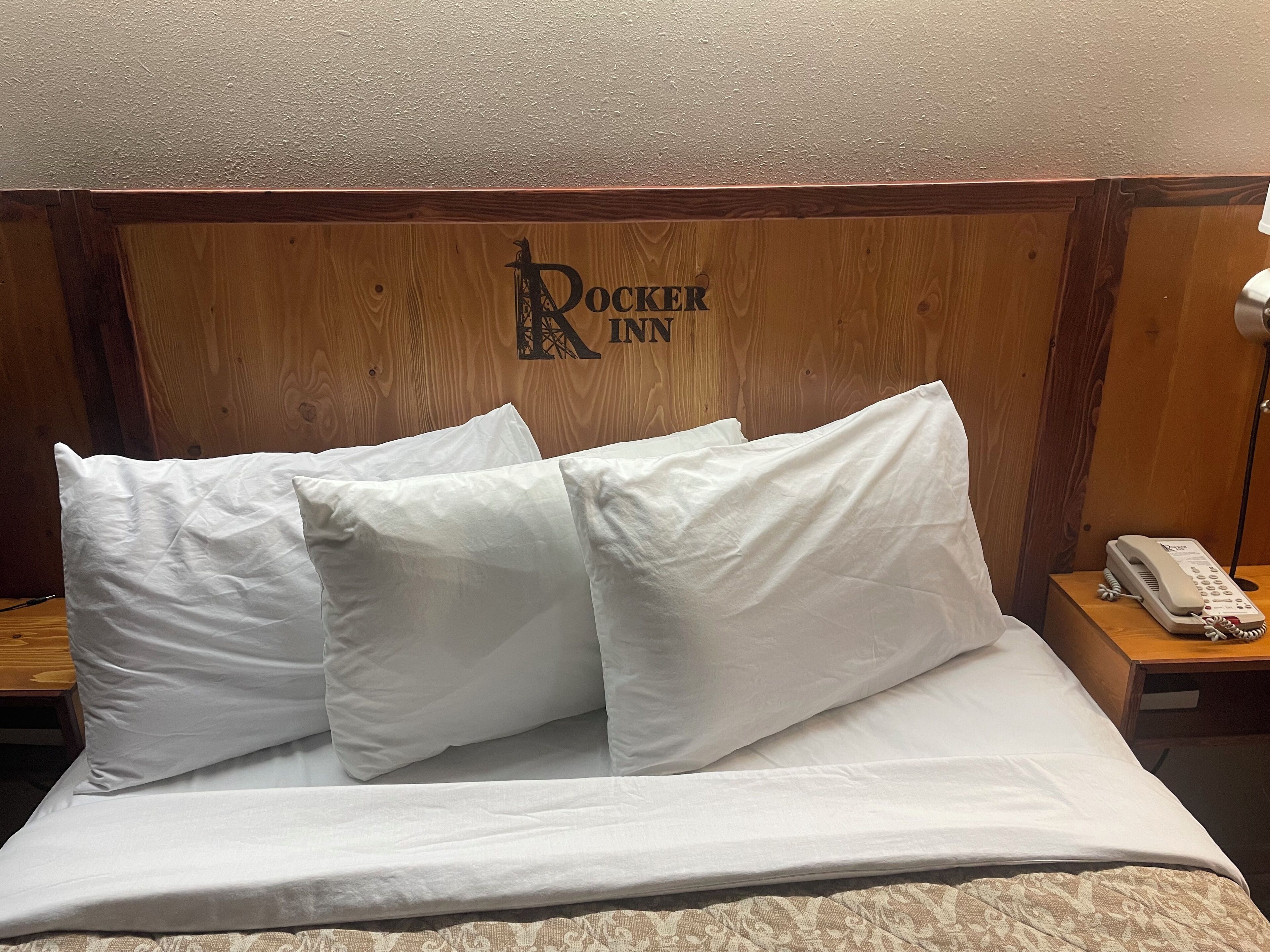 A bed with white linens and pillows, a wooden headboard with the text "DOKKER INN", and a bedside phone on a wooden nightstand.