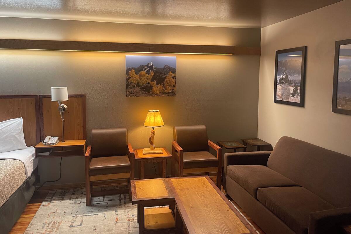 A hotel room with a bed, a desk with a lamp, two chairs, a sofa, and a coffee table. There are framed pictures on the wall.